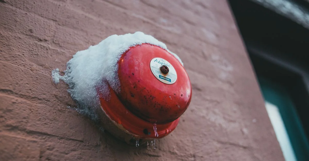 Un bouton d'alarme incendie rouge fixé à un mur en briques, recouvert de neige, mettant en avant la sécurité dans des conditions hivernales.