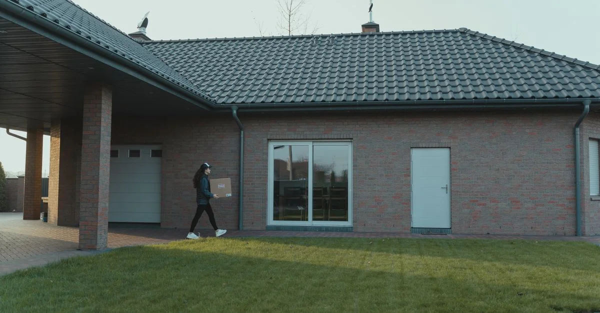 Personne livrant un colis à une maison moderne en briques avec un toit en tuiles et une pelouse verte.
