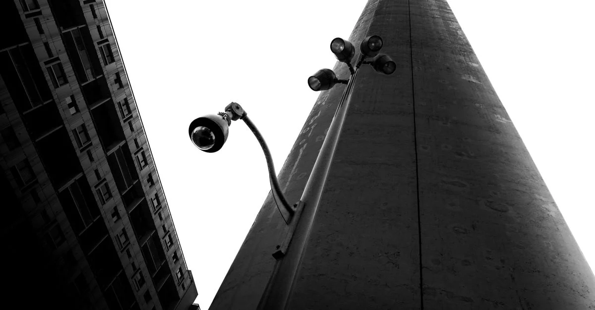 Photo en noir et blanc d'un lampadaire et d'une caméra de sécurité devant un bâtiment moderne et le ciel.